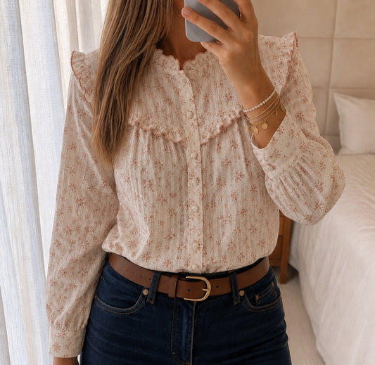 Mujer con blusa Soreya de algodón crudo con estampado floral rosa, cuello alto con volante y canesú con volantes, combinada con vaqueros y cinturón marrón