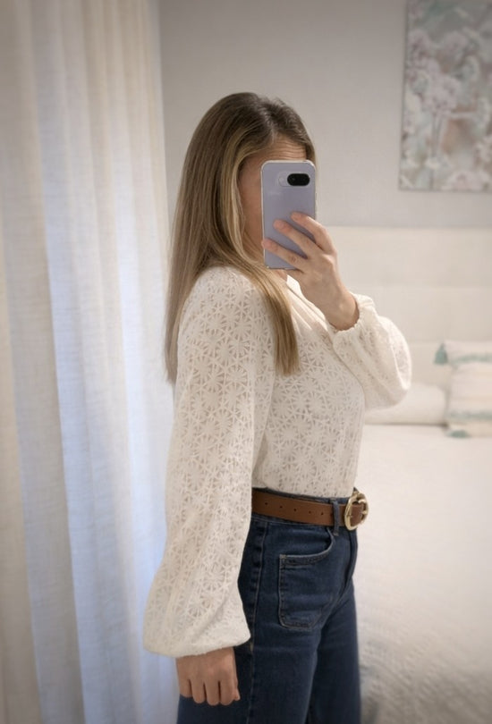 Mujer de perfil con blusa calada color crudo de manga abullonada, combinada con vaqueros y cinturón marrón