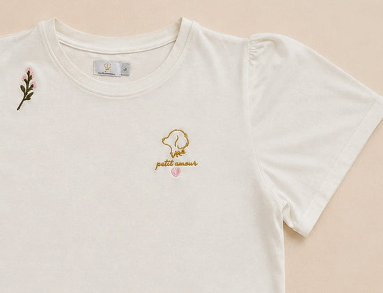 Detalle del bordado a mano de la camiseta Petit Amour Rosé: logo de Leo & Magnolia en hilo dorado, la inscripción petit amour y un pequeño corazón rosa, con una florecilla bordada junto al hombro