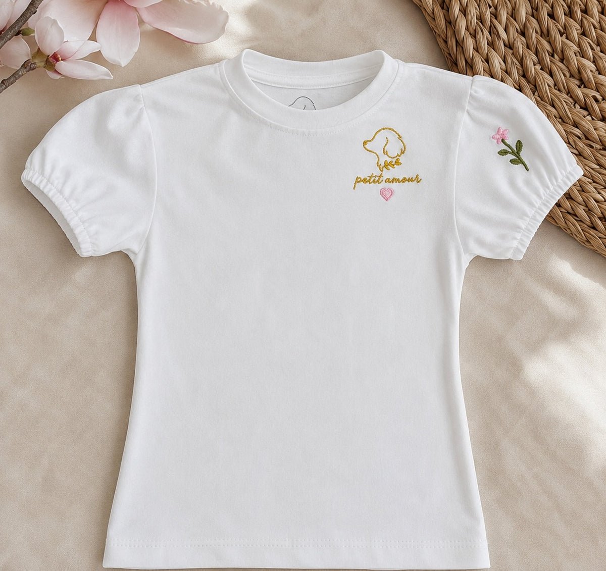 Camiseta Fleur de niña en algodón blanco con mangas abullonadas, bordado dorado petit amour con corazón rosa y una florecilla bordada en la manga, sobre fibra natural y flores de magnolia