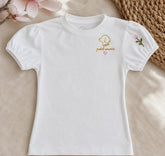 Camiseta Fleur de niña en algodón blanco con mangas abullonadas, bordado dorado petit amour con corazón rosa y una florecilla bordada en la manga, sobre fibra natural y flores de magnolia